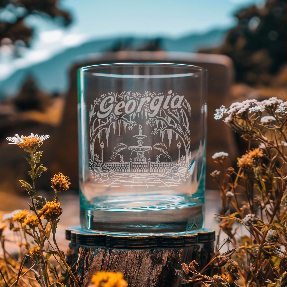 A clear glass rocks tumbler with an etched Georgia state design, displayed on a rustic wooden slice coaster outdoors with wildflowers and a blurred mountain background.