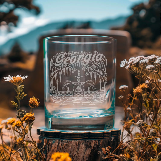 A clear glass rocks tumbler with an etched Georgia state design, displayed on a rustic wooden slice coaster outdoors with wildflowers and a blurred mountain background.