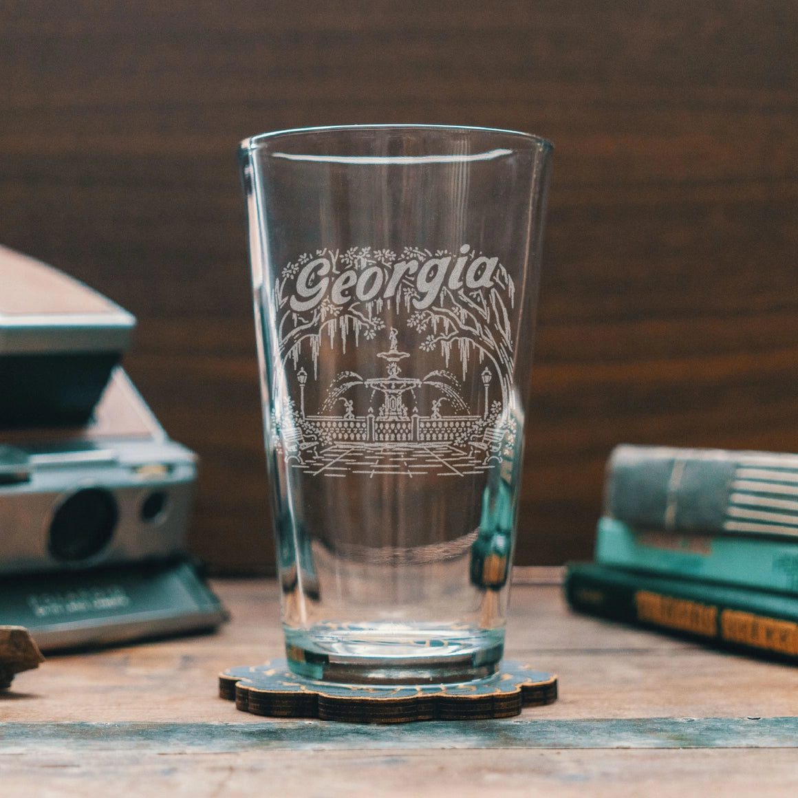 A clear glass pint tumbler with an etched 'Georgia' fountain and bridge design, resting on a wooden coaster on a rustic table with vintage props in the background.