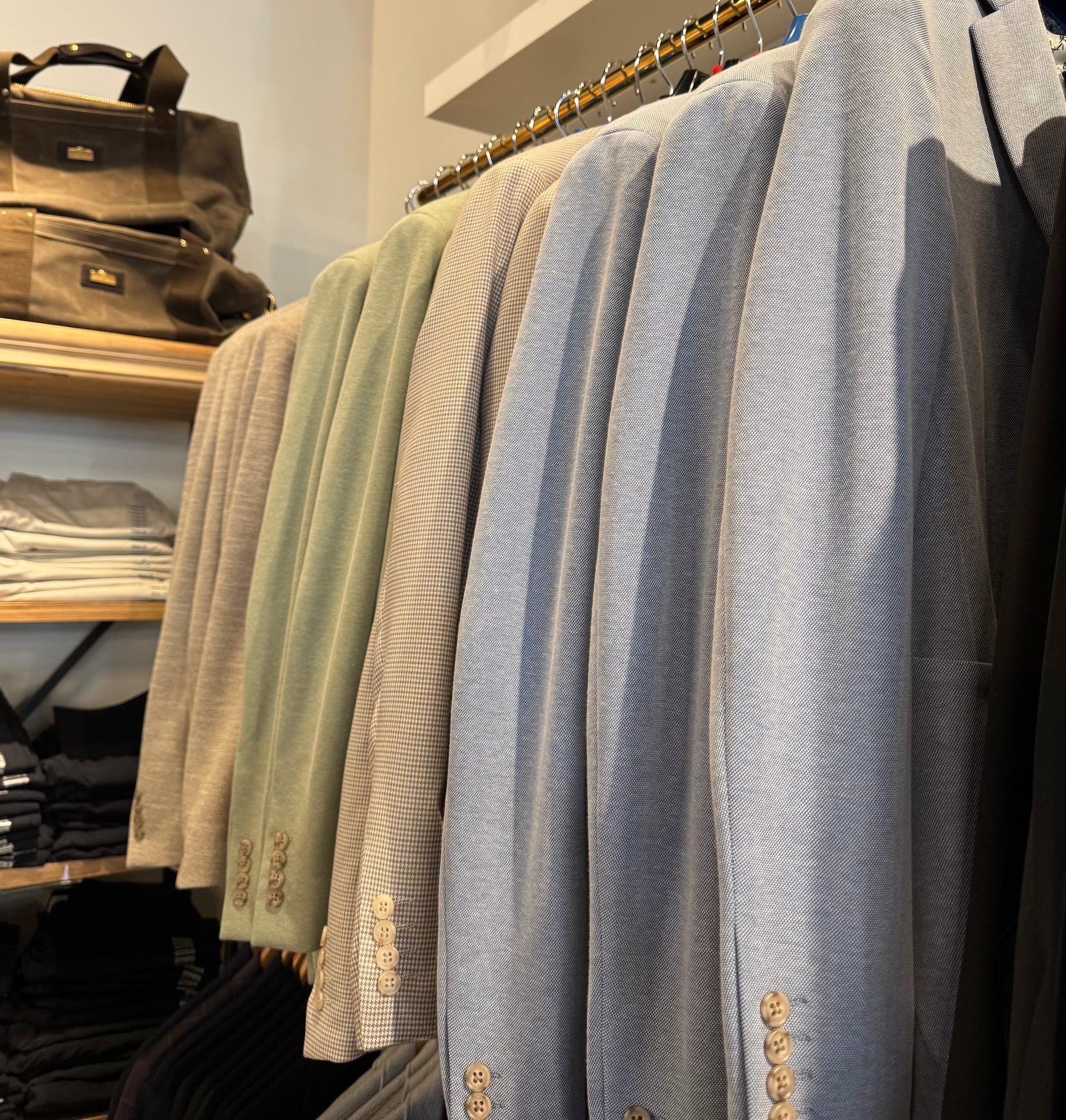 A close-up of a clothing rack displaying a row of tailored blazers in muted colors, including light blue, sage green, beige, and gray, each detailed with gold button accents near the cuffs.
