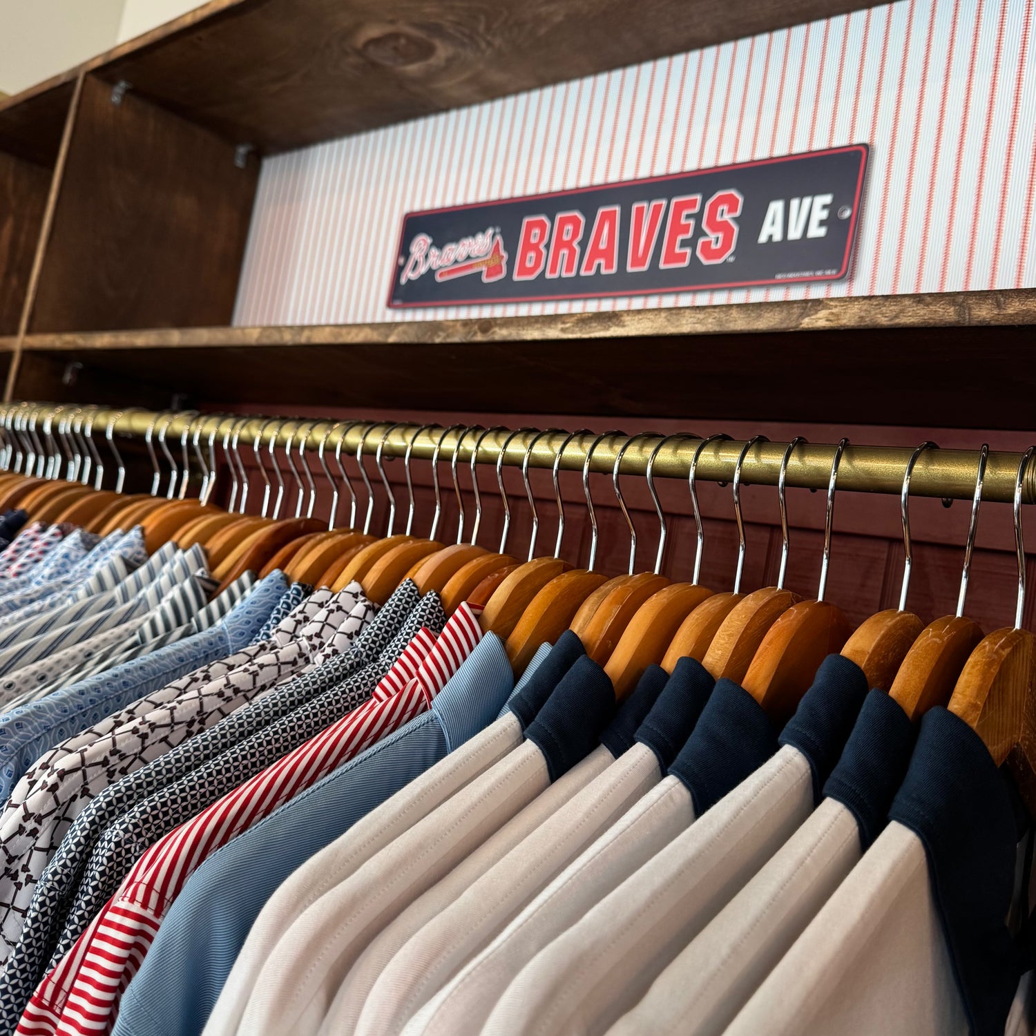 A close-up of a clothing rack inside a wooden closet, showing multiple shirts and tops on wooden hangers. A decorative sign reading "Braves AVE" hangs on the shelf behind the rack.
