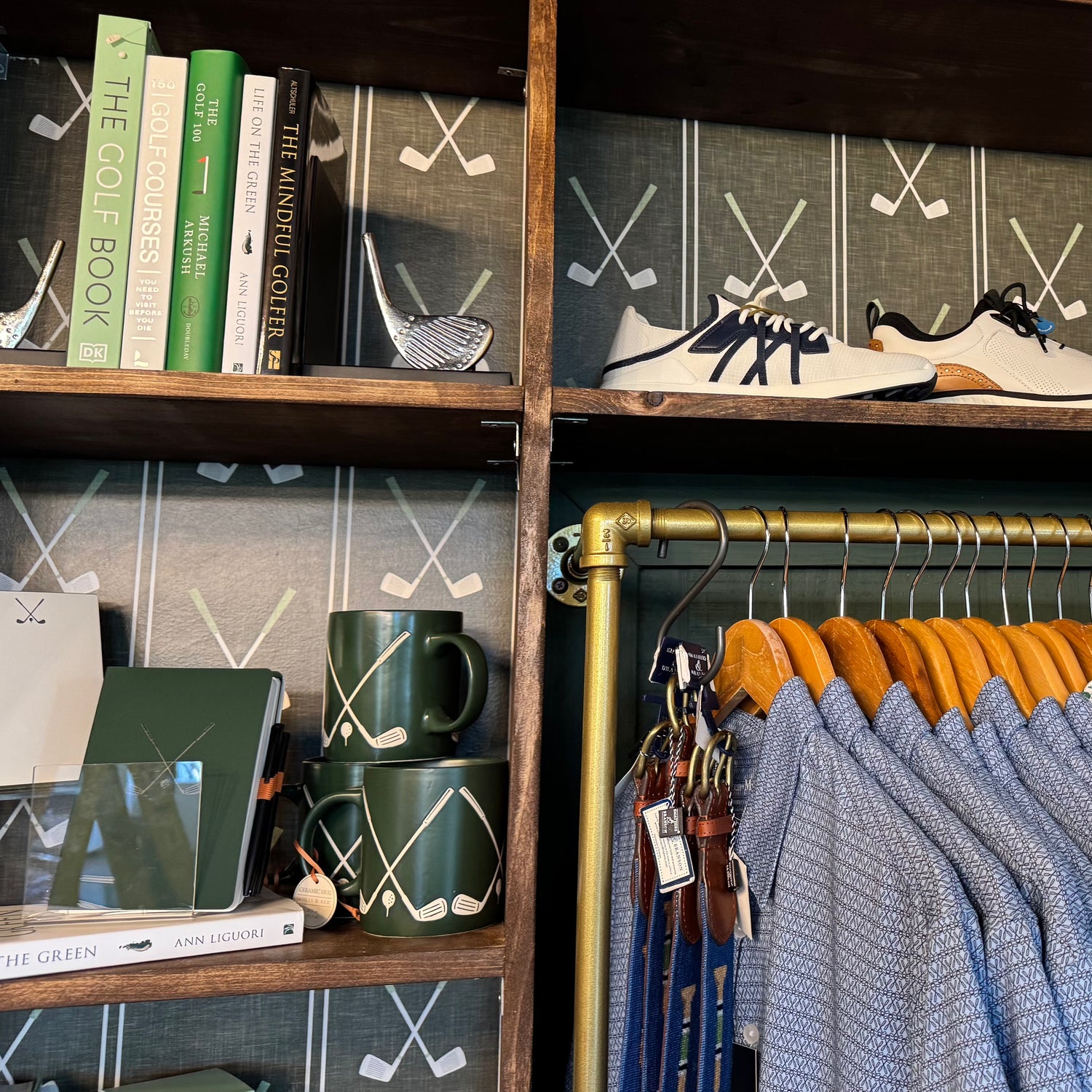 A styled interior shelf and clothing rack area with golf-themed decor and apparel. Visible items include patterned wallpaper with crossed golf clubs, a wooden shelf with books, mugs, and a golf club figurine, a pair of white and navy golf shoes on a shelf, and blue patterned shirts hanging on wooden hangers from a gold pipe rack.