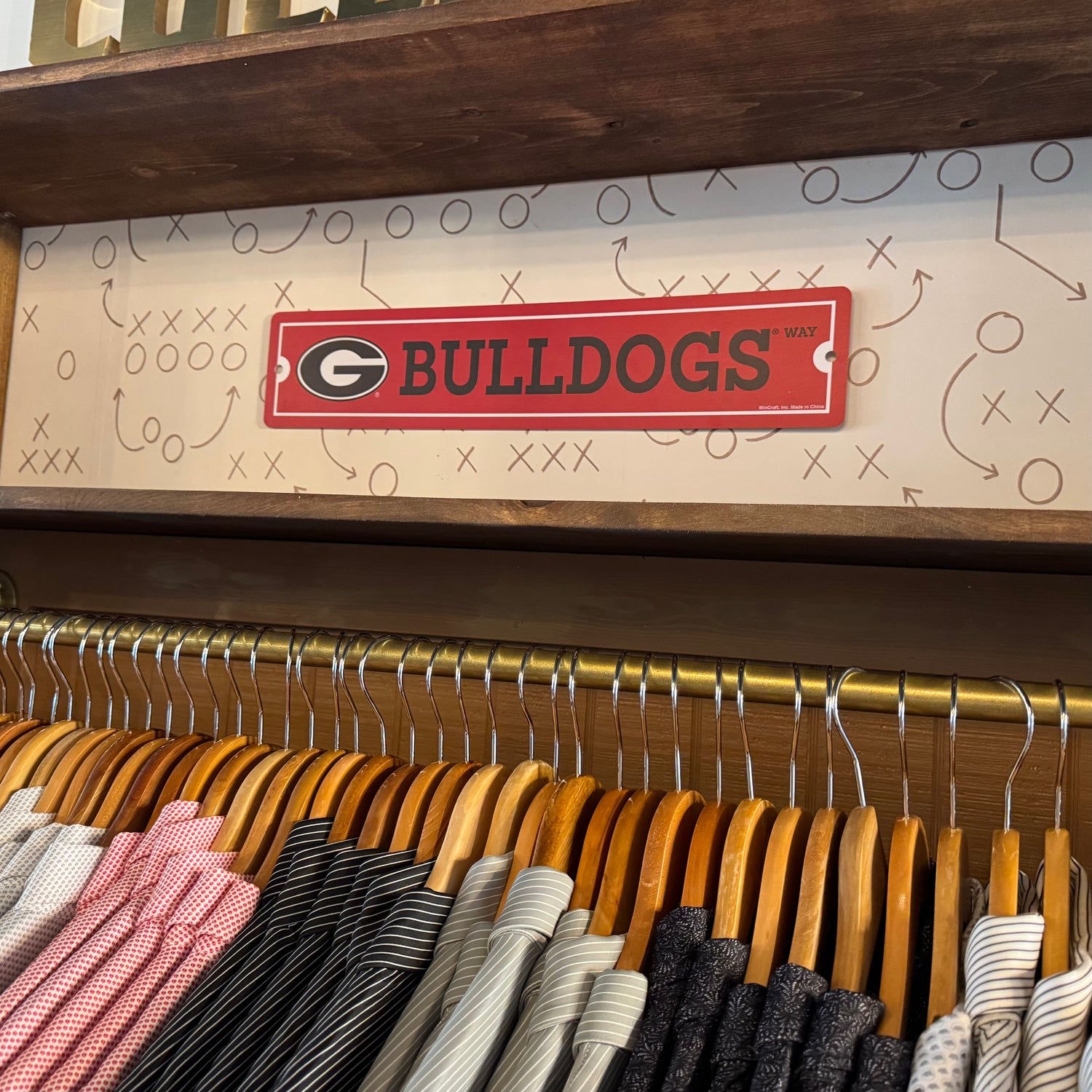 A red rectangular sign with the Georgia Bulldogs logo and wordmark hanging on a beige patterned wall above a clothing rack filled with shirts and ties.